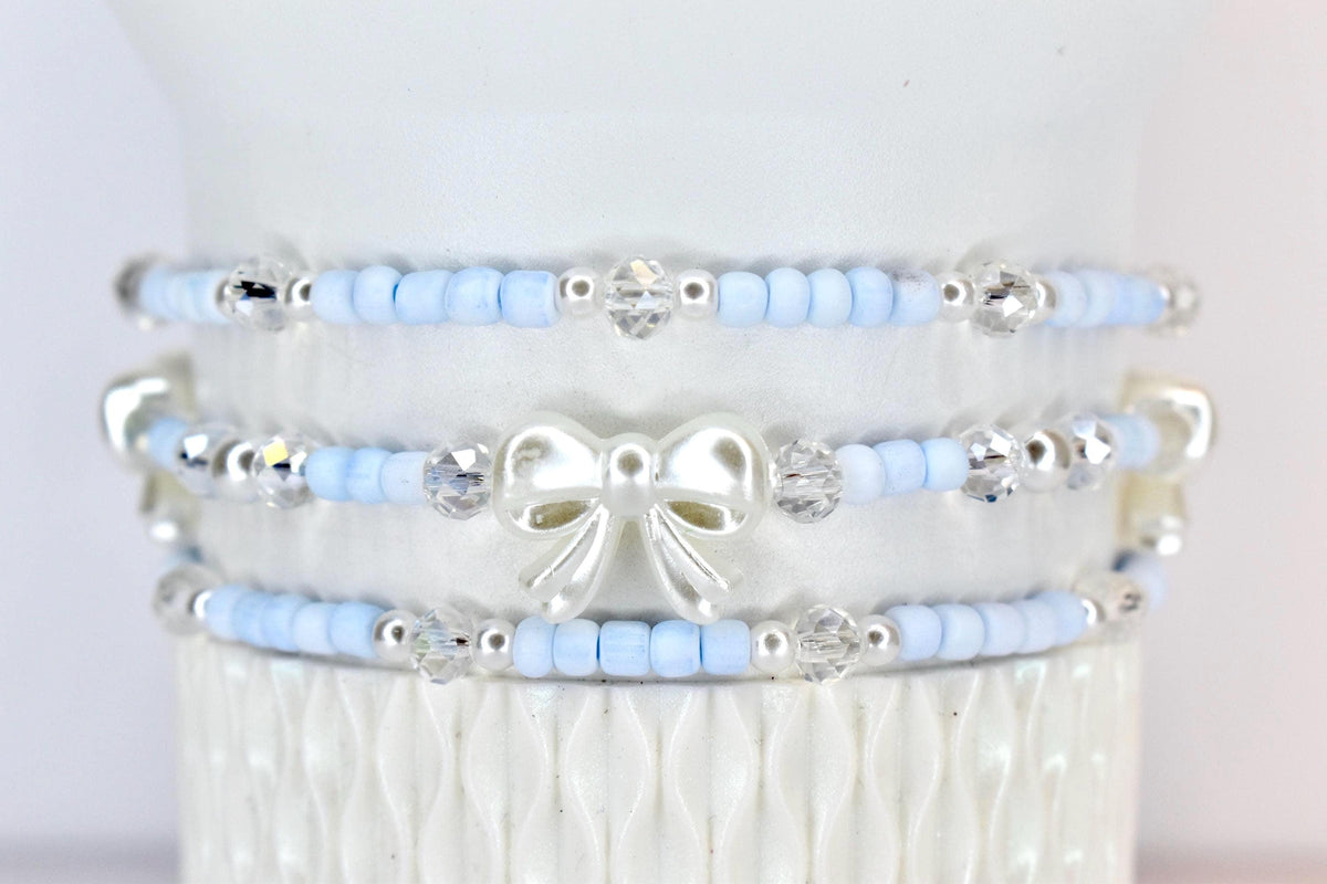 Three beaded bracelets. Each bracelet features light blue beads, silver accents, and clear crystal beads. The middle bracelet has a white bow charm. The bracelets are stacked on a white surface.