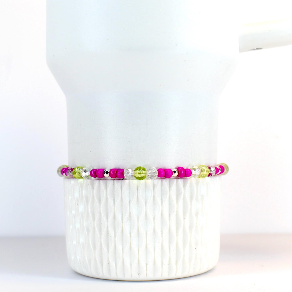 A beaded bracelet featuring a vibrant mix of colors. The bracelet showcases bright pink, clear, and light green beads, interspersed with small silver-colored beads. The bracelet is displayed on a white Stanley tumbler.