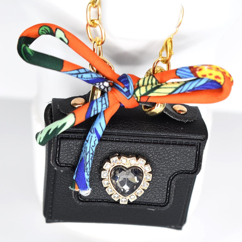 A small, black handbag pendant with a gold chain. The bag features a heart-shaped rhinestone embellishment and is adorned with a colorful orange and patterned scarf tied in a bow. The chain is gold-colored.