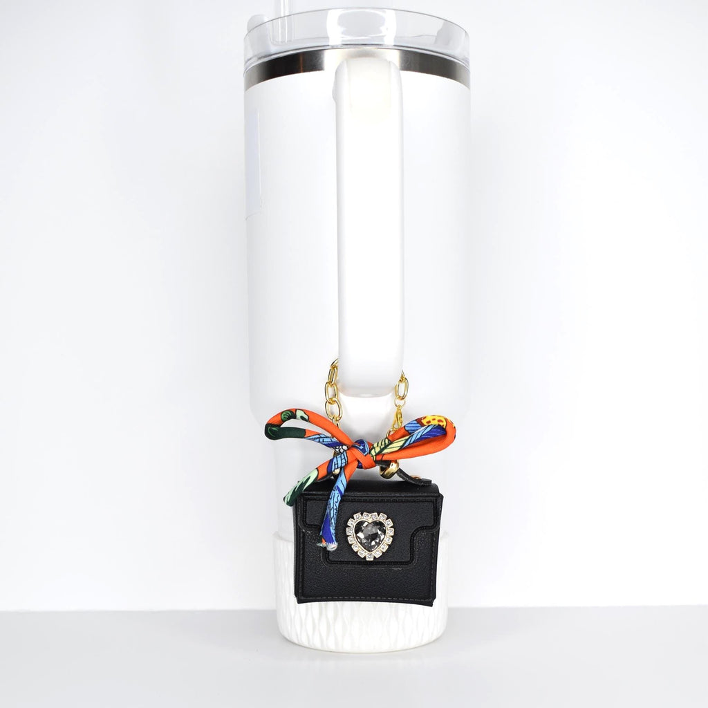 A white tumbler with a handle, adorned with a small black purse pendant. The purse features a rhinestone heart and is attached with a gold chain and a colorful scarf. The tumbler has a clear lid and a straw.