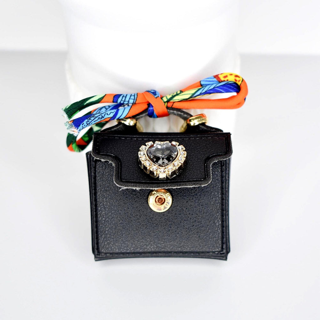 A small, black handbag with a gold clasp and a heart-shaped jewel. The bag has a colorful scarf tied to the handle. The jewel is surrounded by smaller clear stones.