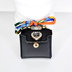 A small, black handbag with a gold clasp and a heart-shaped jewel. The bag has a colorful scarf tied to the handle. The jewel is surrounded by smaller clear stones.