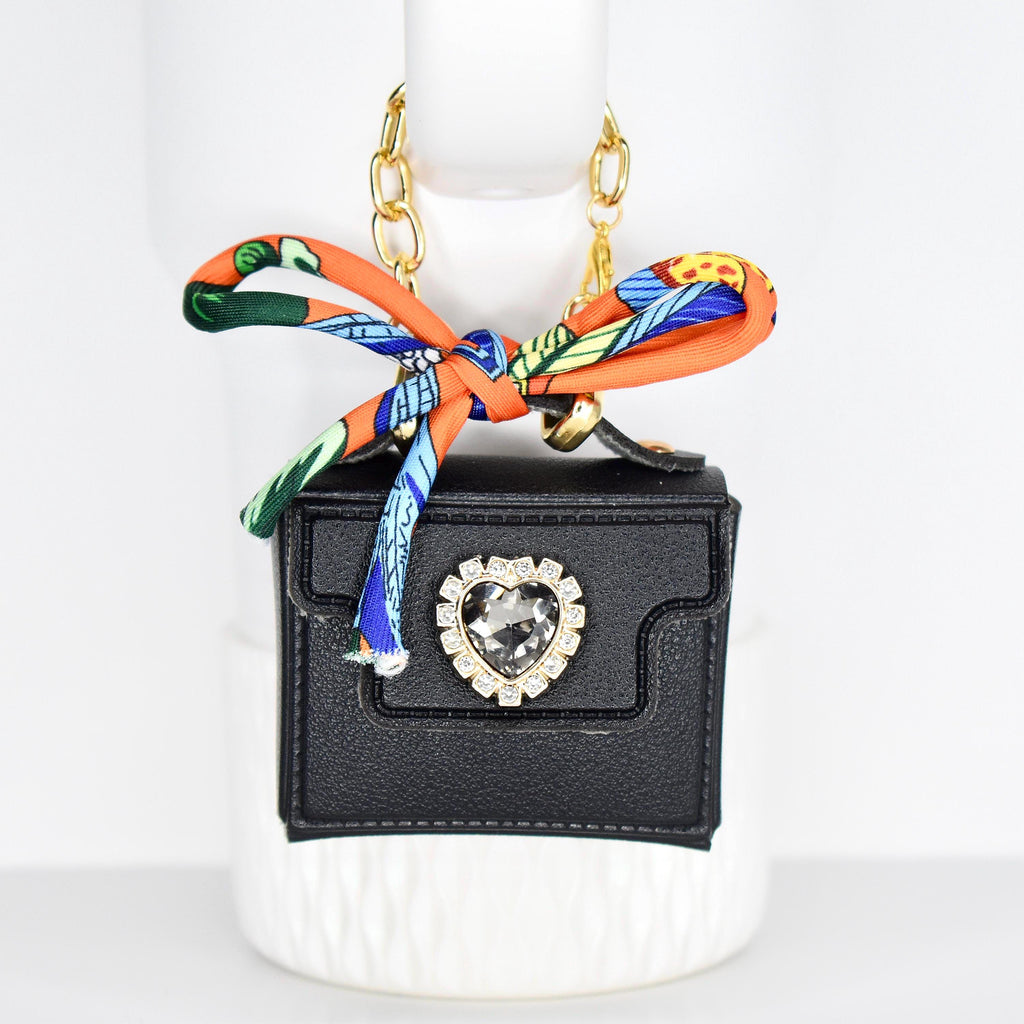 A small, black handbag with a gold chain strap and a decorative bow. The bow is orange with a colorful pattern. The bag features a heart-shaped rhinestone embellishment. The bag is approximately 3 inches wide.