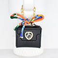 A small, black handbag with a gold chain strap and a decorative bow. The bow is orange with a colorful pattern. The bag features a heart-shaped rhinestone embellishment. The bag is approximately 3 inches wide.