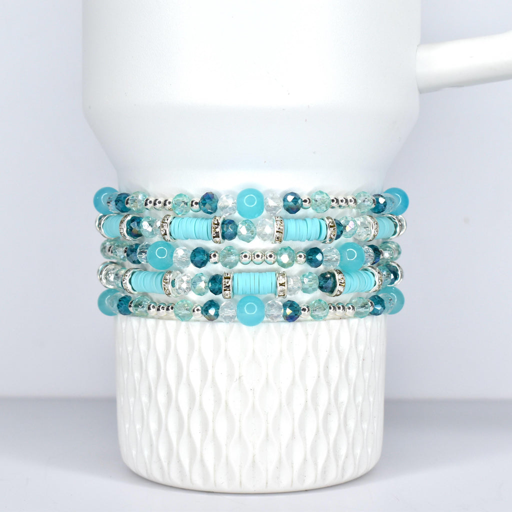Stack of five beaded bracelets in shades of turquoise, teal, and clear. The bracelets feature a mix of round and cylindrical beads, with silver-toned accents. The bracelets are displayed on a white, Stanley tumbler.