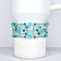 Stack of five beaded bracelets in shades of turquoise, teal, and clear. The bracelets feature a mix of round and cylindrical beads, with silver-toned accents. The bracelets are displayed on a white, Stanley tumbler.