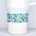 Stack of five beaded bracelets in shades of turquoise, teal, and clear. The bracelets feature a mix of round and cylindrical beads, with silver-toned accents. The bracelets are displayed on a white, Stanley tumbler.