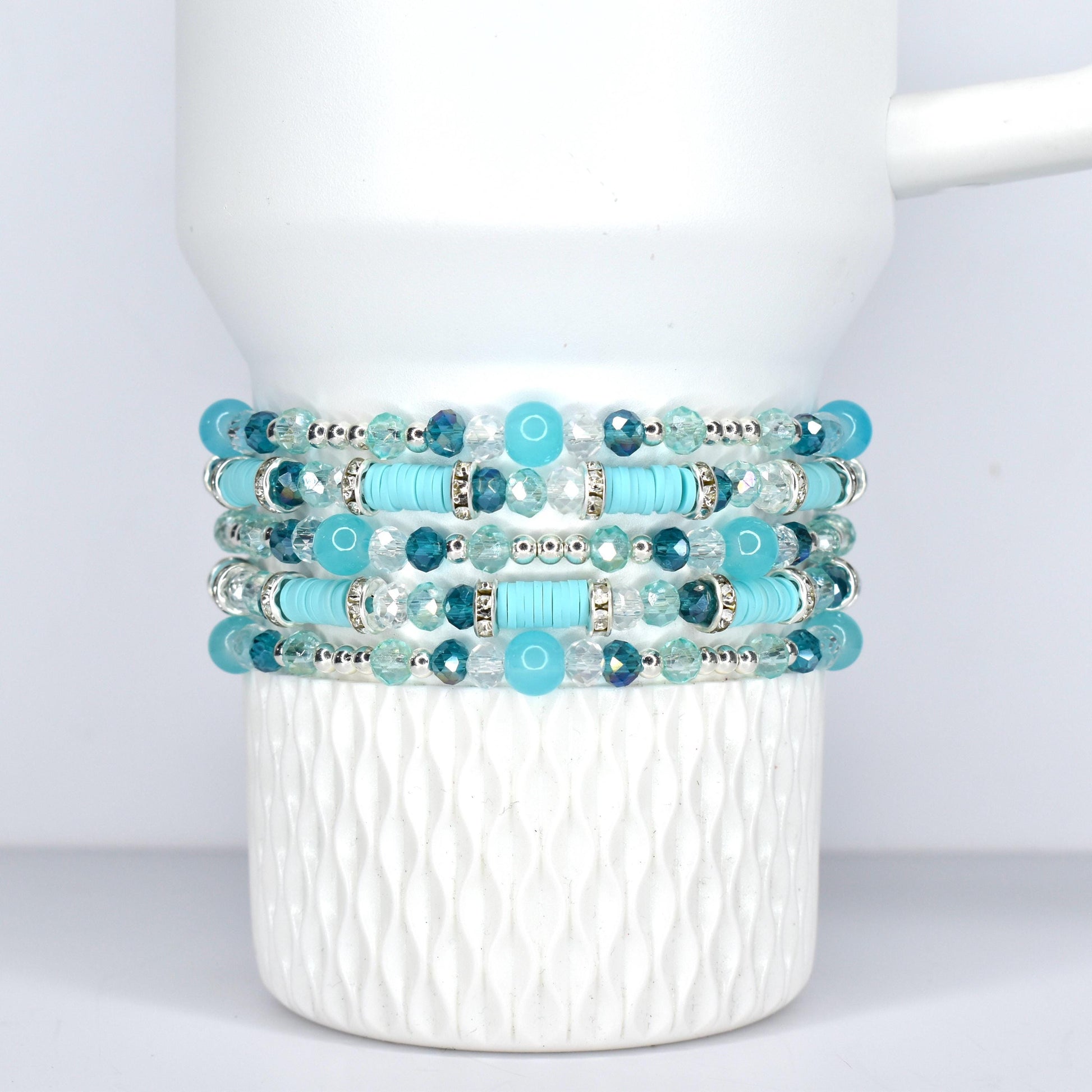 Stack of five beaded bracelets in shades of turquoise, teal, and clear. The bracelets feature a mix of round and cylindrical beads, with silver-toned accents. The bracelets are displayed on a white, Stanley tumbler.