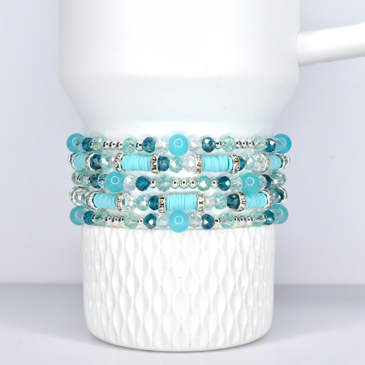 Stack of five beaded bracelets in shades of turquoise, teal, and clear. The bracelets feature a mix of round and cylindrical beads, with silver-toned accents. The bracelets are displayed on a white, Stanley tumbler.