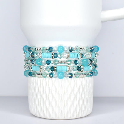 Stack of five beaded bracelets in shades of turquoise, teal, and clear. The bracelets feature a mix of round and cylindrical beads, with silver-toned accents. The bracelets are displayed on a white, Stanley tumbler.