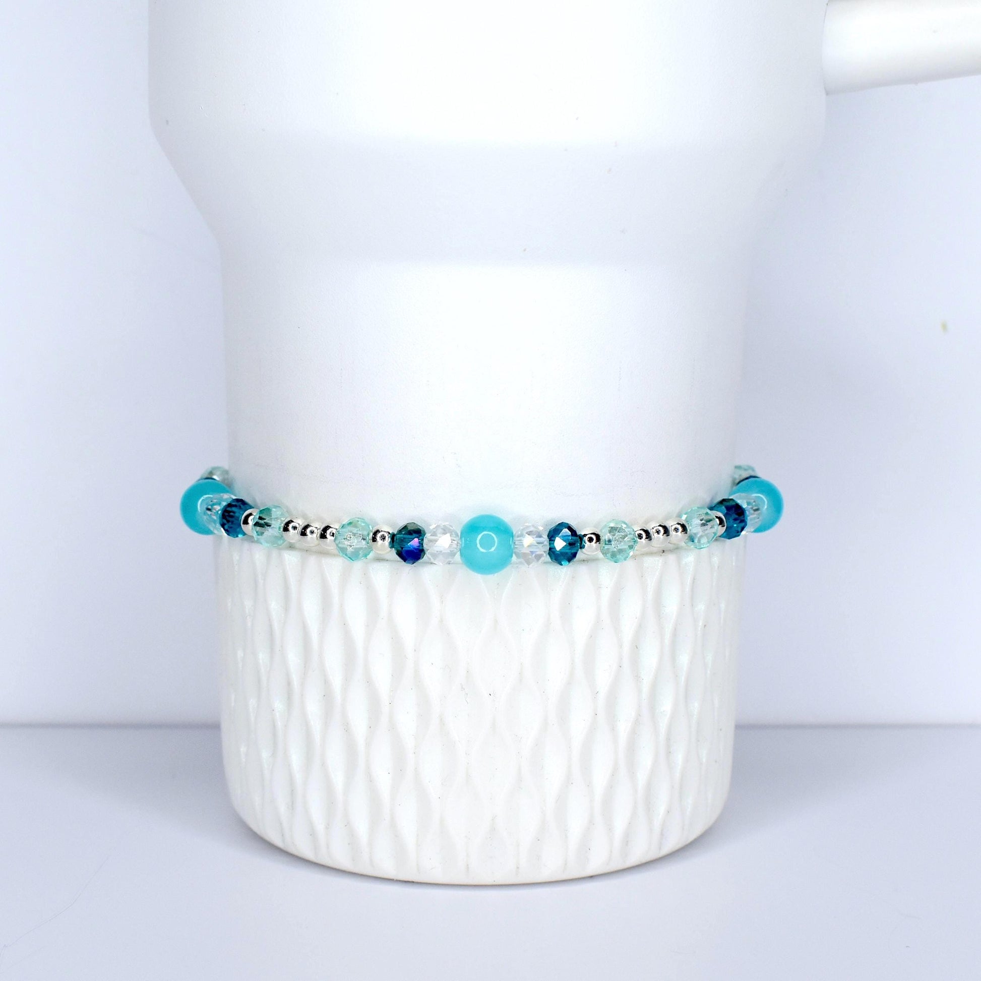 A beaded bracelet with a mix of blue and clear beads. The bracelet features various shades of blue, including turquoise and navy, interspersed with clear, faceted beads. The beads are strung on a silver-colored chain. The bracelet is displayed on a white, textured surface.