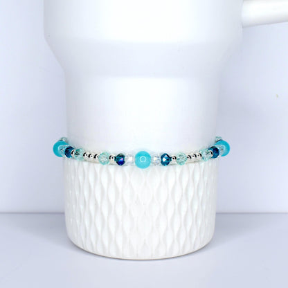 A beaded bracelet with a mix of blue and clear beads. The bracelet features various shades of blue, including turquoise and navy, interspersed with clear, faceted beads. The beads are strung on a silver-colored chain. The bracelet is displayed on a white, textured surface.