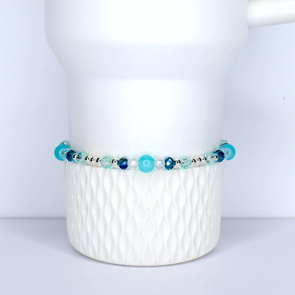 A beaded bracelet with a mix of blue and clear beads. The bracelet features various shades of blue, including turquoise and navy, interspersed with clear, faceted beads. The beads are strung on a silver-colored chain. The bracelet is displayed on a white, textured surface.