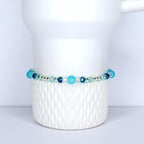 A beaded bracelet with a mix of blue and clear beads. The bracelet features various shades of blue, including turquoise and navy, interspersed with clear, faceted beads. The beads are strung on a silver-colored chain. The bracelet is displayed on a white, textured surface.