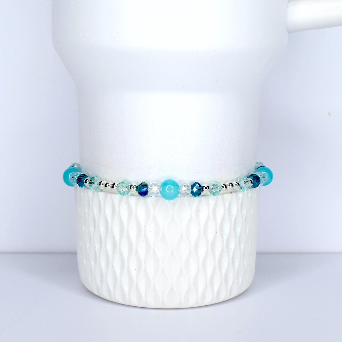 A beaded bracelet with a mix of blue and clear beads. The bracelet features various shades of blue, including turquoise and navy, interspersed with clear, faceted beads. The beads are strung on a silver-colored chain. The bracelet is displayed on a white, textured surface.