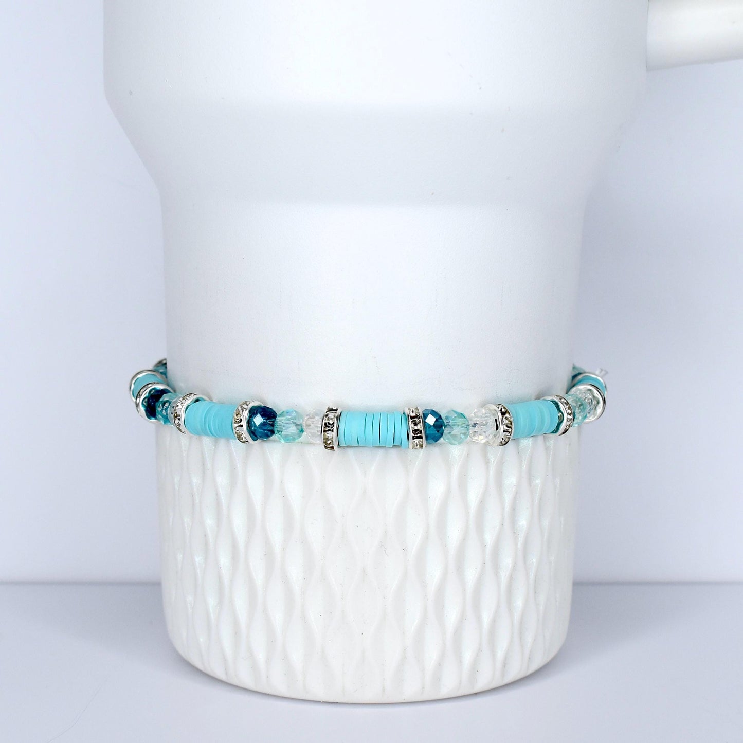 A turquoise beaded bracelet with silver accents and clear and blue crystal beads. The bracelet is displayed on a white, textured ceramic surface. The bracelet features a mix of round and flat beads.