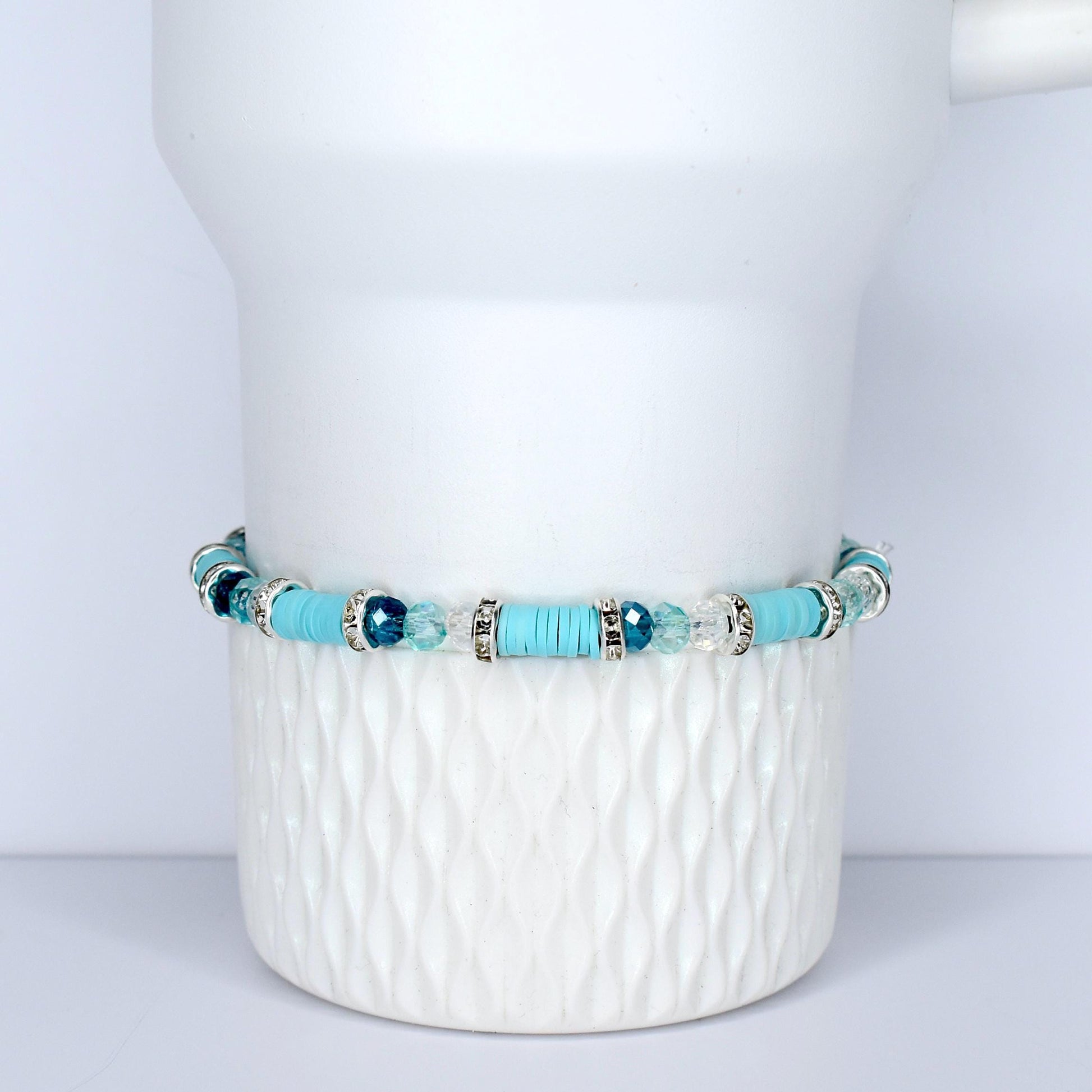 A turquoise beaded bracelet with silver accents and clear and blue crystal beads. The bracelet is displayed on a white, textured ceramic surface. The bracelet features a mix of round and flat beads.
