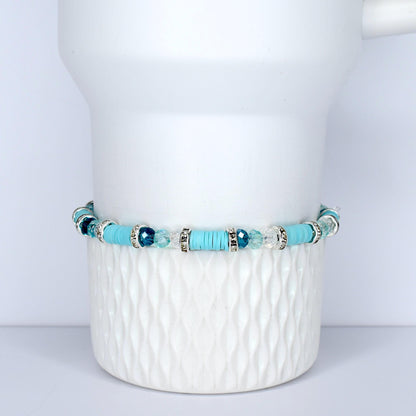 A turquoise beaded bracelet with silver accents and clear and blue crystal beads. The bracelet is displayed on a white, textured ceramic surface. The bracelet features a mix of round and flat beads.