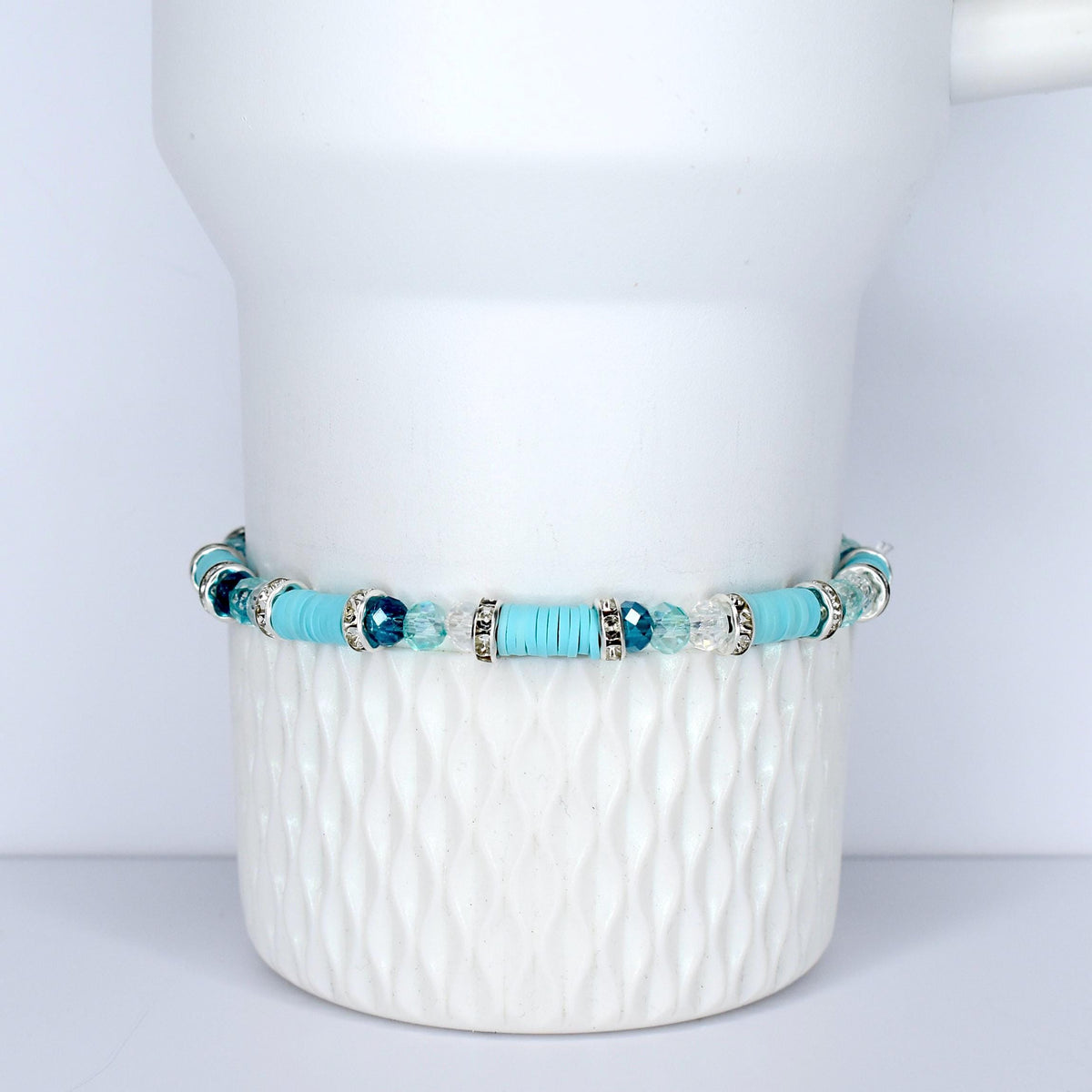 A turquoise beaded bracelet with silver accents and clear and blue crystal beads. The bracelet is displayed on a white, textured ceramic surface. The bracelet features a mix of round and flat beads.
