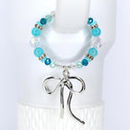 A beaded bracelet featuring turquoise and clear beads, accented with silver-toned spacers. A silver bow charm dangles from the center. The bracelet is displayed against a white background.