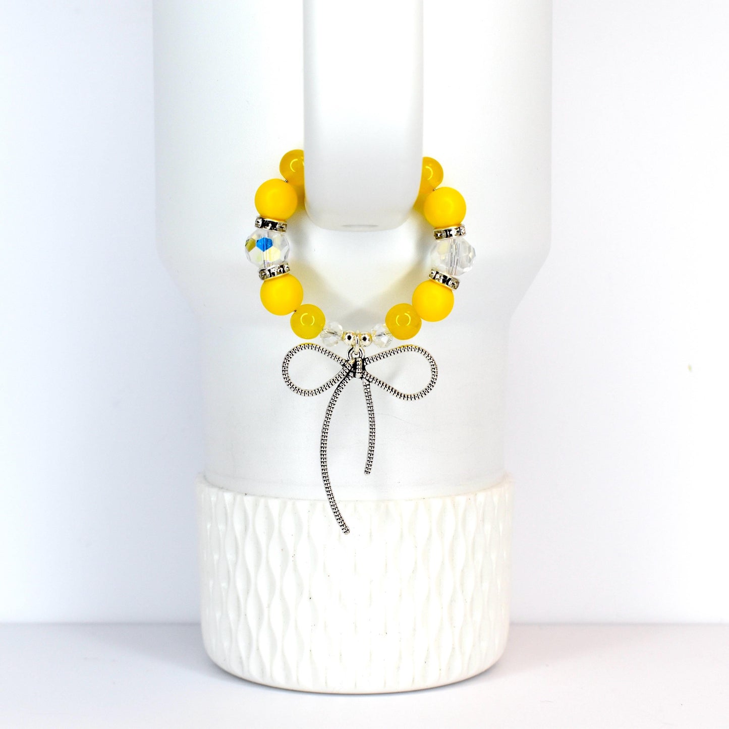 A yellow beaded bracelet with a silver bow charm. The bracelet features large, round yellow beads interspersed with clear, sparkling beads. The bow charm hangs below the bracelet, adding a touch of elegance.