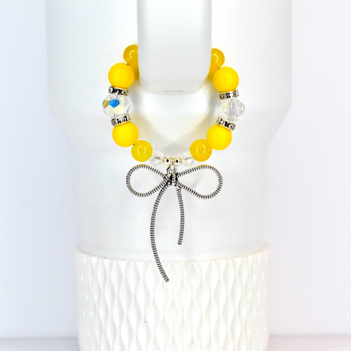 A yellow beaded tumbler handle bracelet with a silver bow charm. The bracelet features large, round yellow beads interspersed with clear, faceted beads. The silver bow charm hangs from the center of the bracelet, adding a touch of elegance.