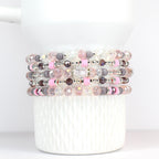 Stack of beaded bracelets in shades of pink, gray, and clear. The bracelets feature a variety of beads, including faceted crystal, round gray beads, and pink spacers. The bracelets are stacked on a white, textured surface.