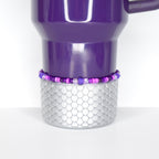 A purple travel mug with a silver, scale-patterned base. A beaded bracelet in shades of purple and pink encircles the base. The mug has a handle and a shiny finish, set against a white background.