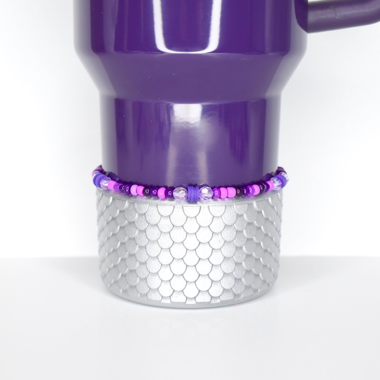 A purple travel mug with a silver, scale-patterned base. A beaded bracelet in shades of purple and pink encircles the base. The mug has a handle and a shiny finish, set against a white background.