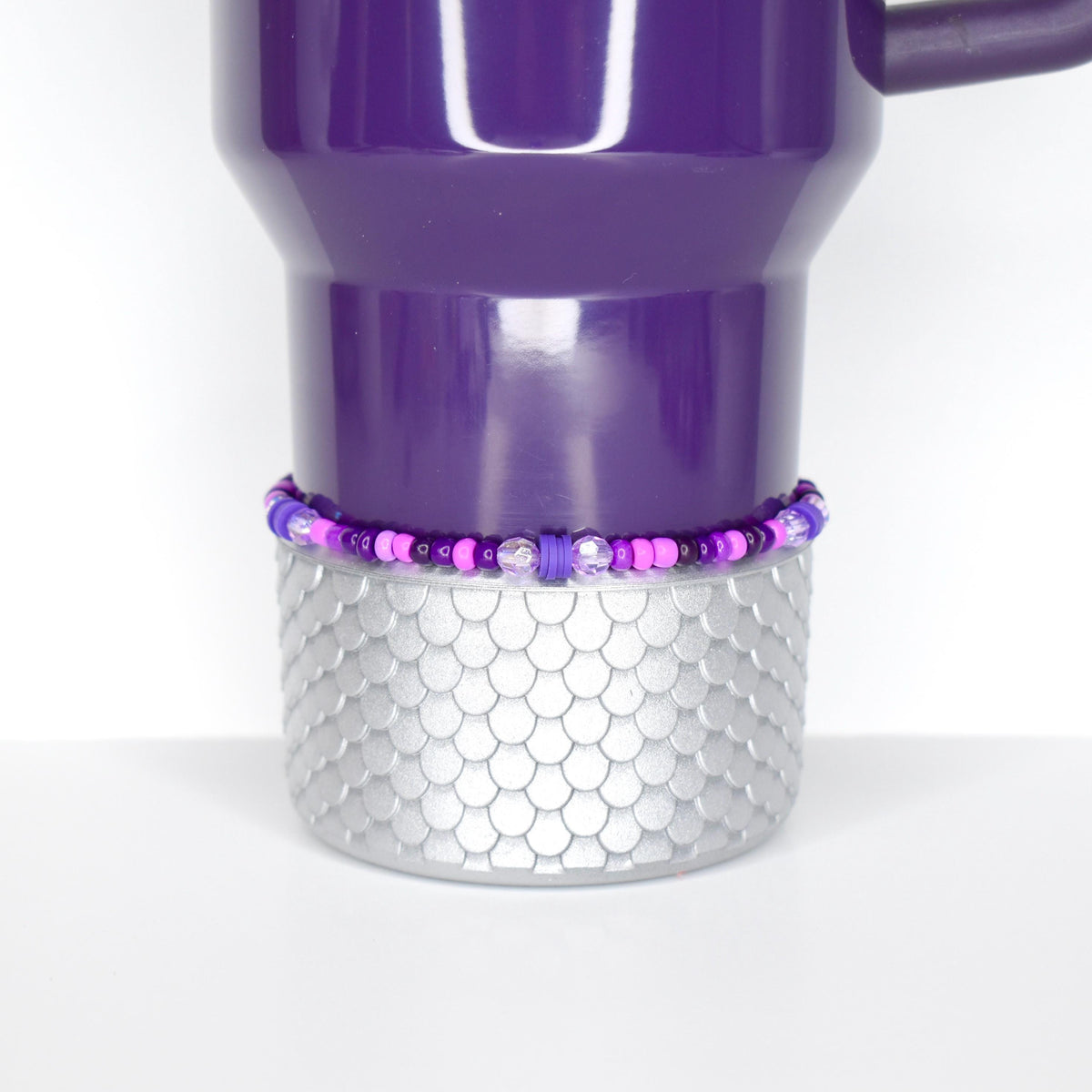 A purple travel mug with a silver, scale-patterned base. A beaded bracelet in shades of purple and pink encircles the base. The mug has a handle and a shiny finish, set against a white background.