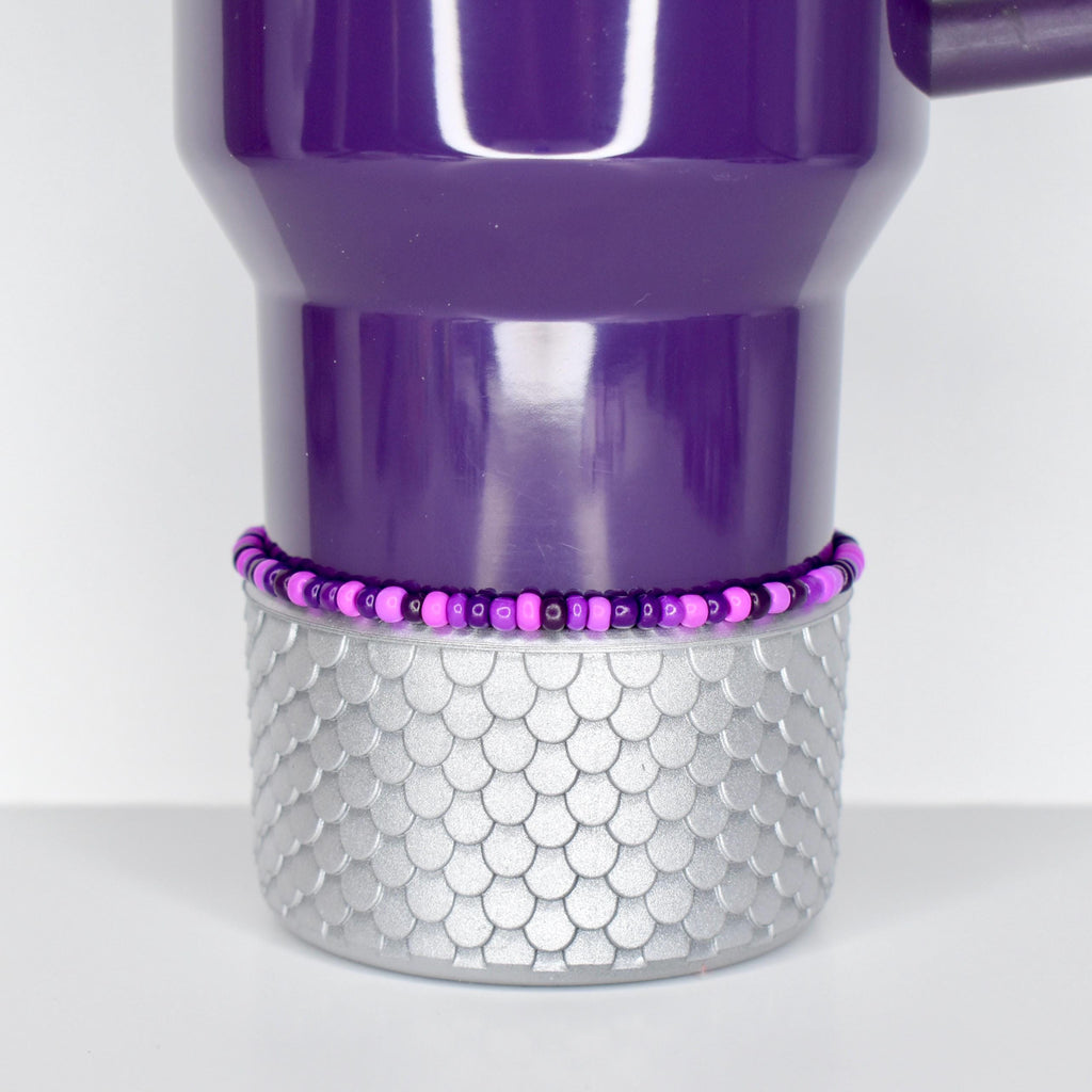 A purple tumbler with a silver, scale-patterned base. A beaded bracelet in shades of purple and pink encircles the tumbler. The tumbler has a handle and a curved top.