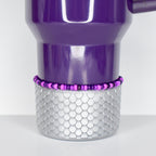 A purple tumbler with a silver, scale-patterned base. A beaded bracelet in shades of purple and pink encircles the tumbler. The tumbler has a handle and a curved top.