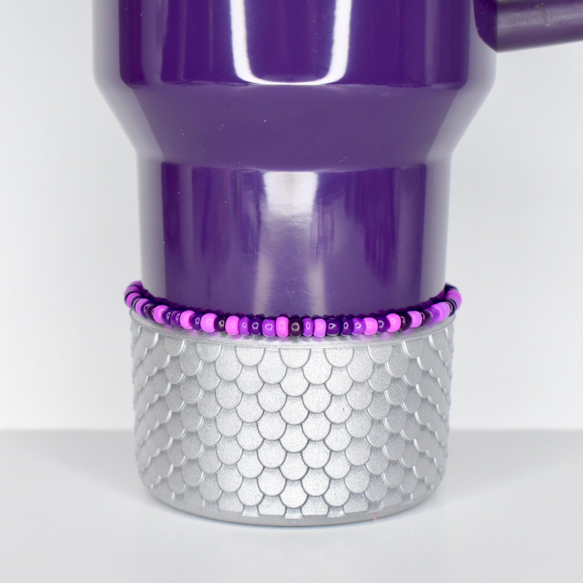 A purple tumbler with a silver, scale-patterned base. A beaded bracelet in shades of purple and pink encircles the tumbler. The tumbler has a handle and a curved top.