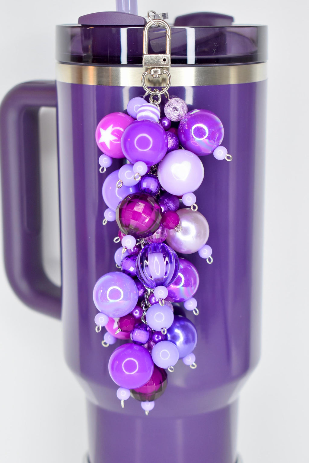 A plum stanley with a silver rim and handle, adorned with a beaded charm. The charm features various shades of purple beads, including round, star-shaped, and faceted designs, attached to a silver keychain.