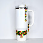 A white insulated tumbler with a handle, a clear lid, and a straw. The tumbler is adorned with beaded charms in red, yellow, green, and black, and gold-colored pendants. Several bracelets with similar colors are wrapped around the base.