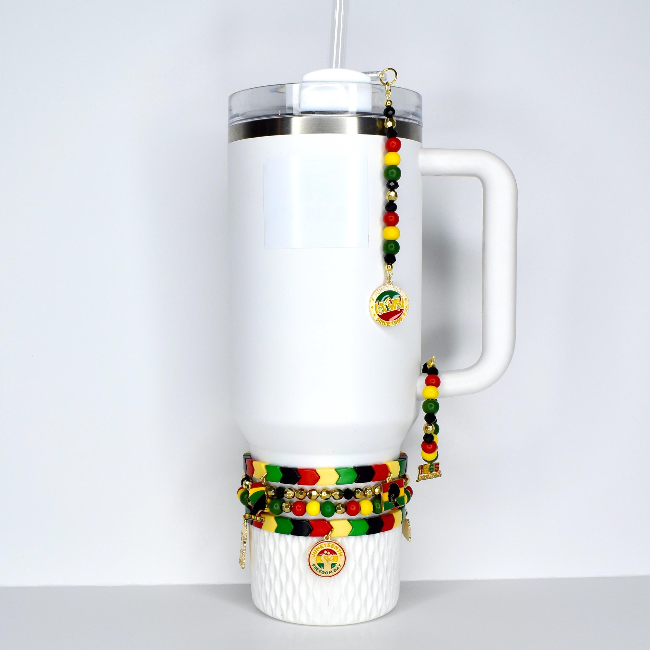 A white insulated tumbler with a handle, a clear lid, and a straw. The tumbler is adorned with beaded charms in red, yellow, green, and black, along with gold-colored pendants. Several bracelets with similar color schemes are wrapped around the base.