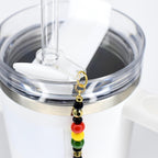 A white tumbler with a clear straw and a Juneteenth charm. The charm features black, gold, red, yellow, and green beads. The tumbler has a silver-colored band around the top.