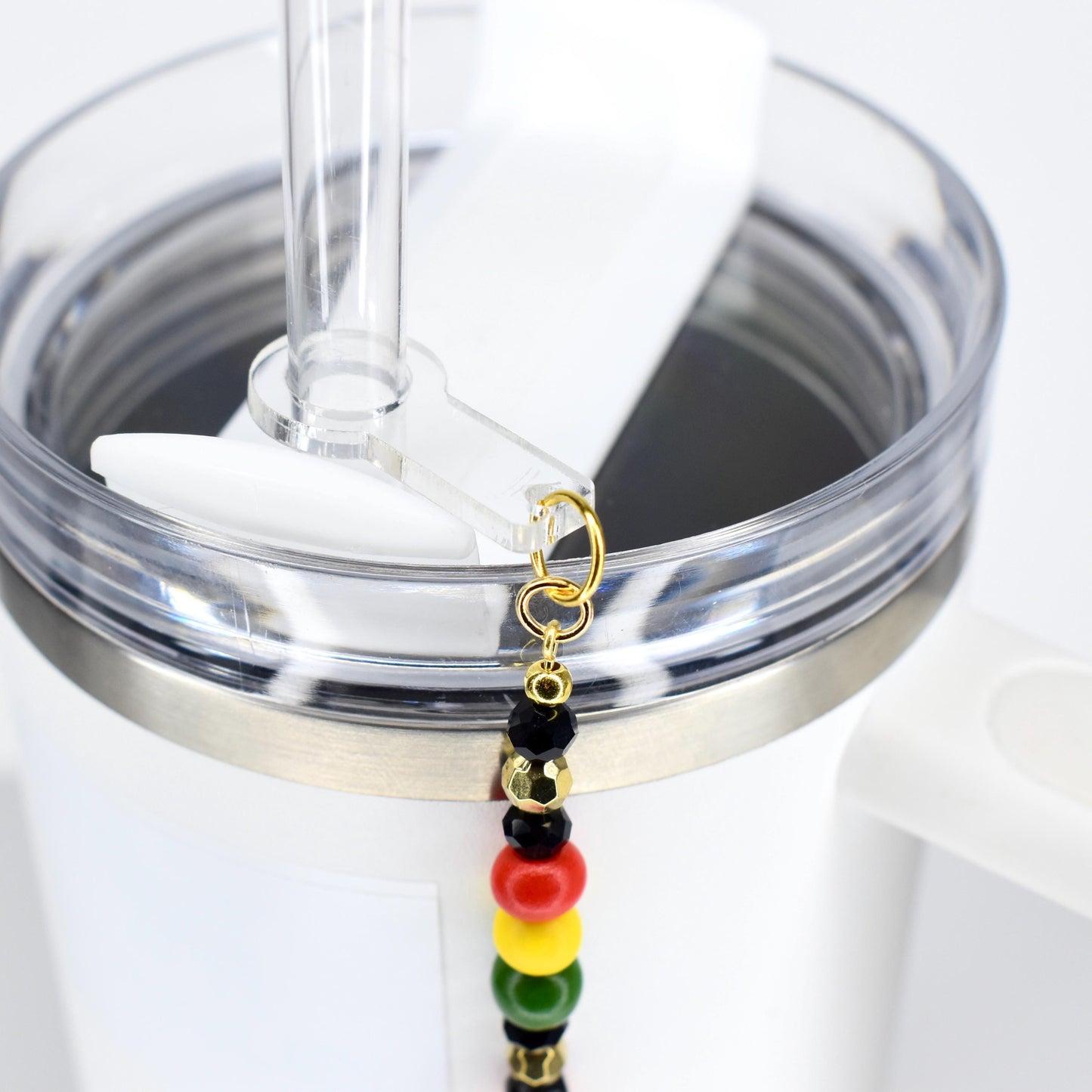 A white tumbler with a clear straw and a Juneteenth charm. The charm features black, gold, red, yellow, and green beads. The tumbler has a silver-colored band around the top.