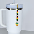 A white Stanley tumbler with a handle and a straw, featuring a colorful beaded Juneteenth celebration charm. The charm has red, yellow, green, and black beads, and a gold-colored pendant with text that reads "Since 1865".