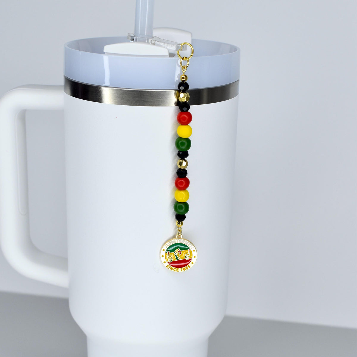 A white Stanley tumbler with a handle and a straw, featuring a colorful beaded Juneteenth celebration charm. The charm has red, yellow, green, and black beads, and a gold-colored pendant with text that reads "Since 1865".