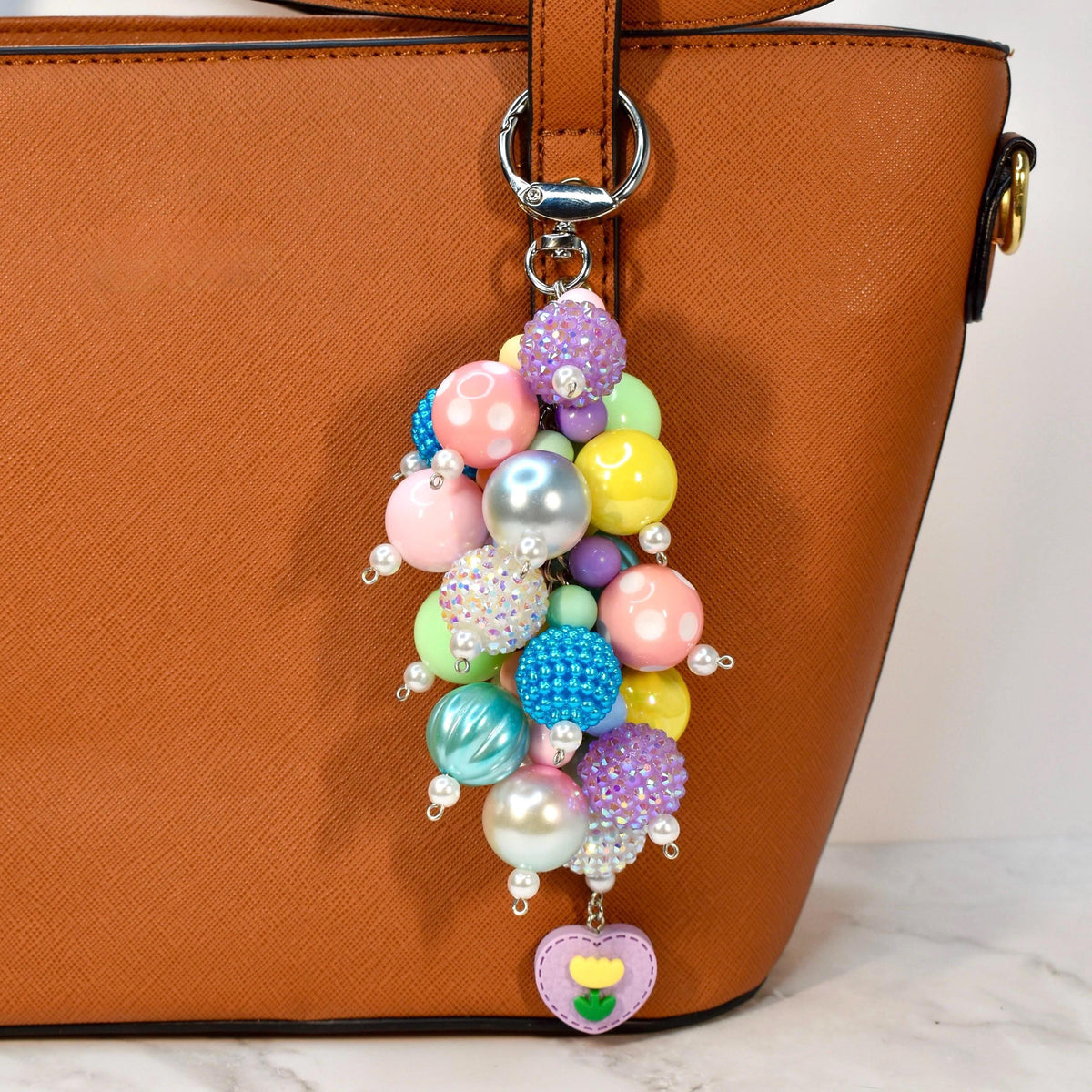 Colorful pastel beaded purse charm hanging from a brown handbag, designed with mixed round beads, pearls, and a heart-shaped floral pendant. Playful spring bag charm and statement keychain accessory, perfect for adding charm to totes and purses.