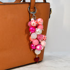 A beaded keychain with a variety of pink, white, and red beads. The keychain has a silver clasp and a heart-shaped charm at the bottom. The beads are different sizes and textures, including some with glitter and heart patterns.