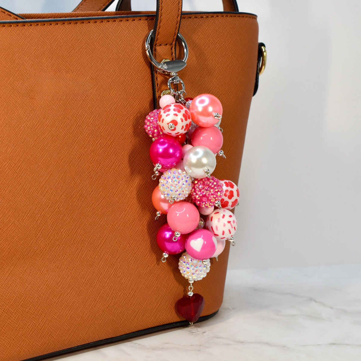 A beaded keychain with a variety of pink, white, and red beads. The keychain has a silver clasp and a heart-shaped charm at the bottom. The beads are different sizes and textures, including some with glitter and heart patterns.