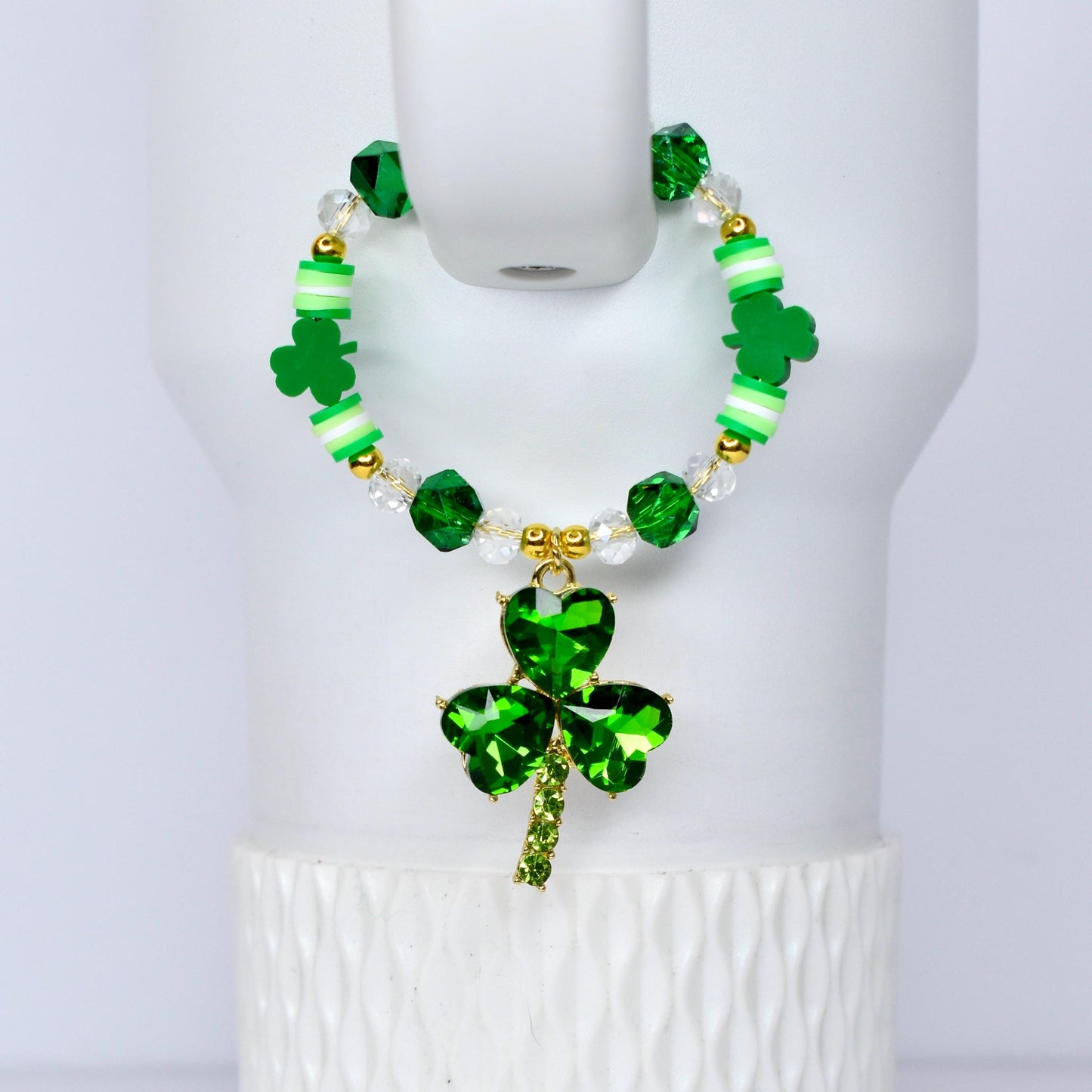 A green beaded handle charm with a shamrock pendant. The bracelet features green, white, and gold beads, with a large, sparkling shamrock charm. The shamrock pendant is made of green crystals and has a gold-colored stem.