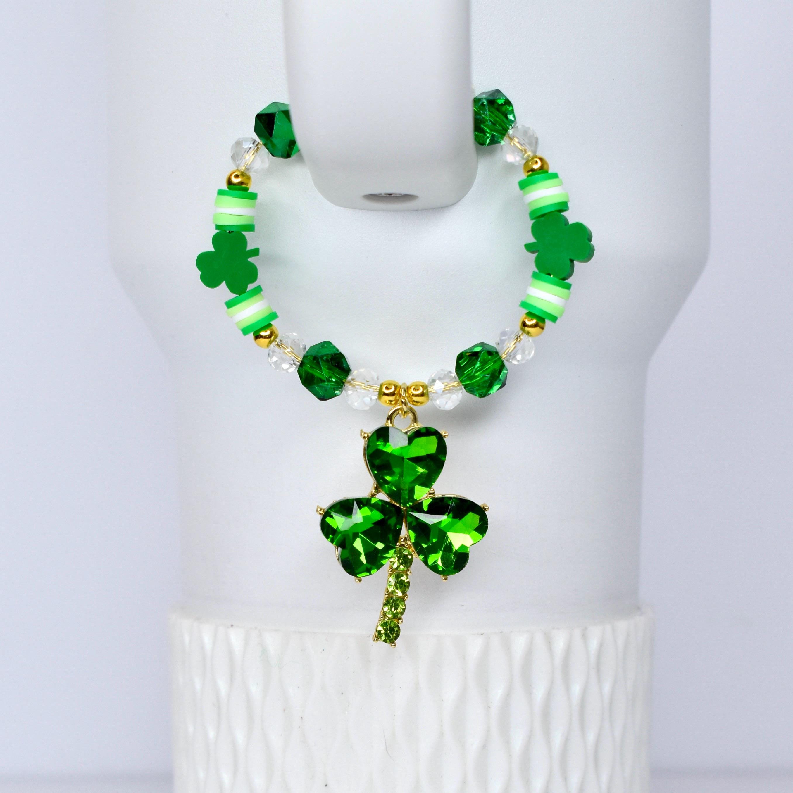 A green beaded handle charm with a shamrock pendant. The bracelet features green, white, and gold beads, with a large, sparkling shamrock charm. The shamrock pendant is made of green crystals and has a gold-colored stem.
