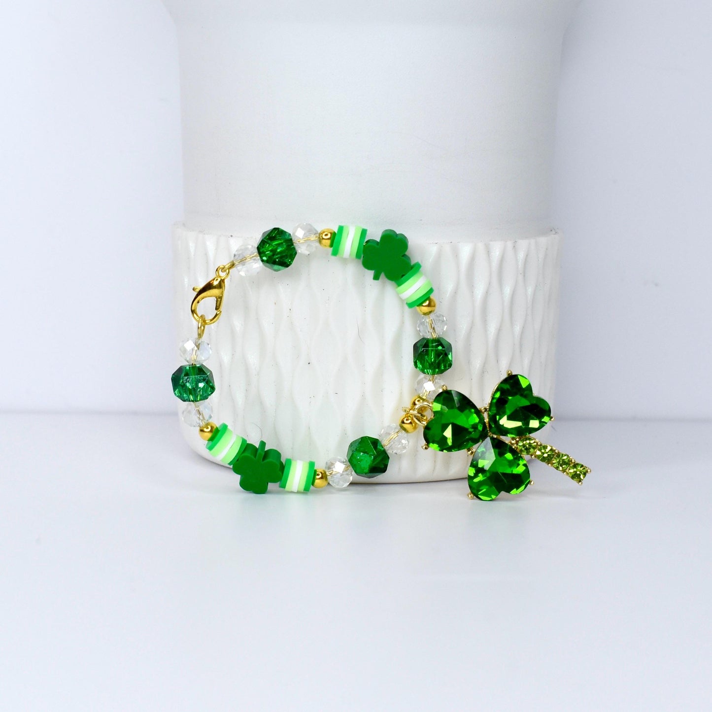 A St. Patrick's Day bracelet featuring green shamrock charms, beads, and accents. The bracelet includes clear and green faceted beads, white and green striped beads, and a large shamrock pendant with a gold clasp.