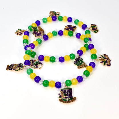 Three strands of Mardi Gras beads with green, yellow, and purple beads. Each strand has decorative charms, including a top hat, a fleur-de-lis, and a beer mug. The beads are on a white background.