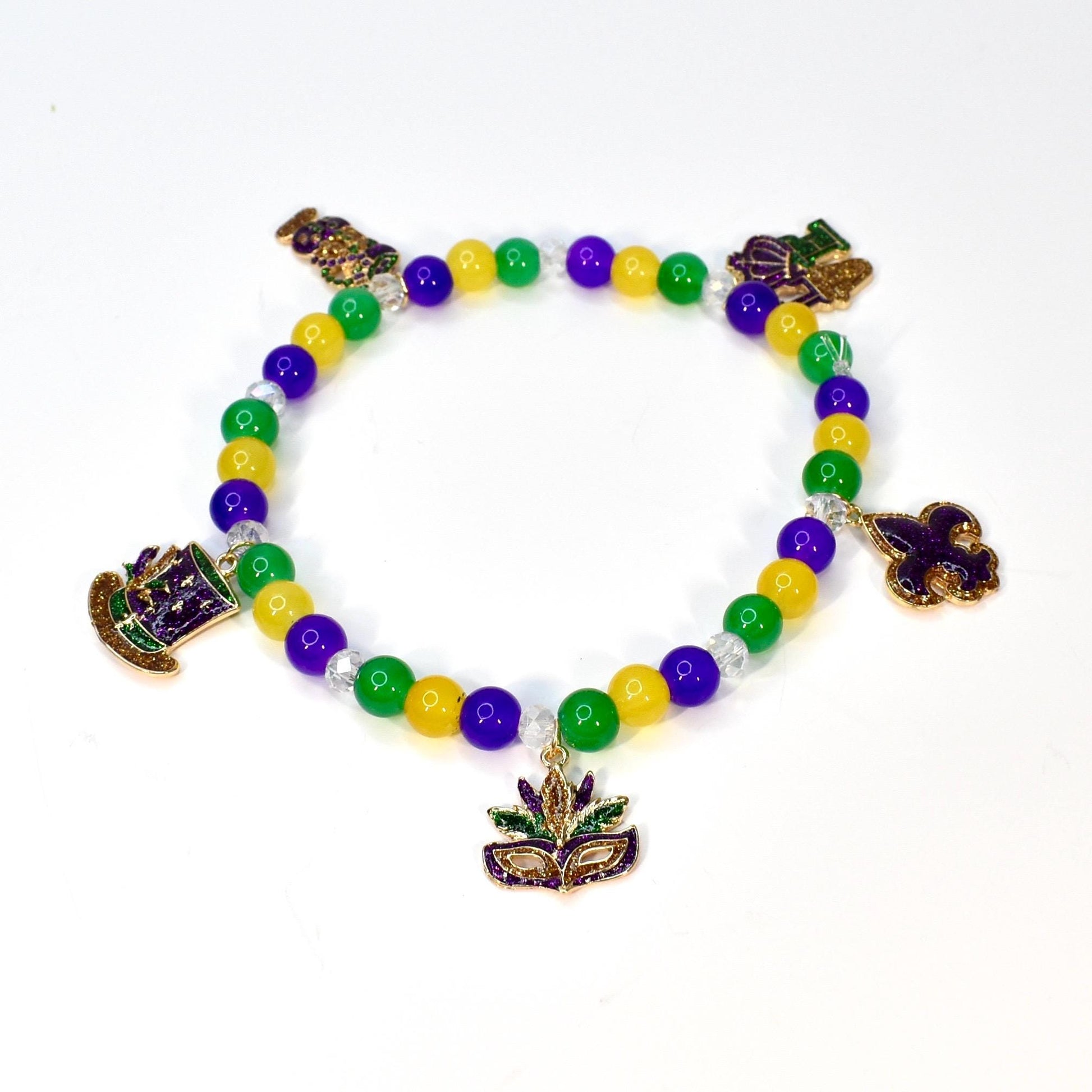 A beaded bracelet with yellow, green, and purple beads. The bracelet features gold-colored charms of a hat, a mask, and a fleur-de-lis. The beads are strung on a clear elastic cord, creating a festive accessory.
