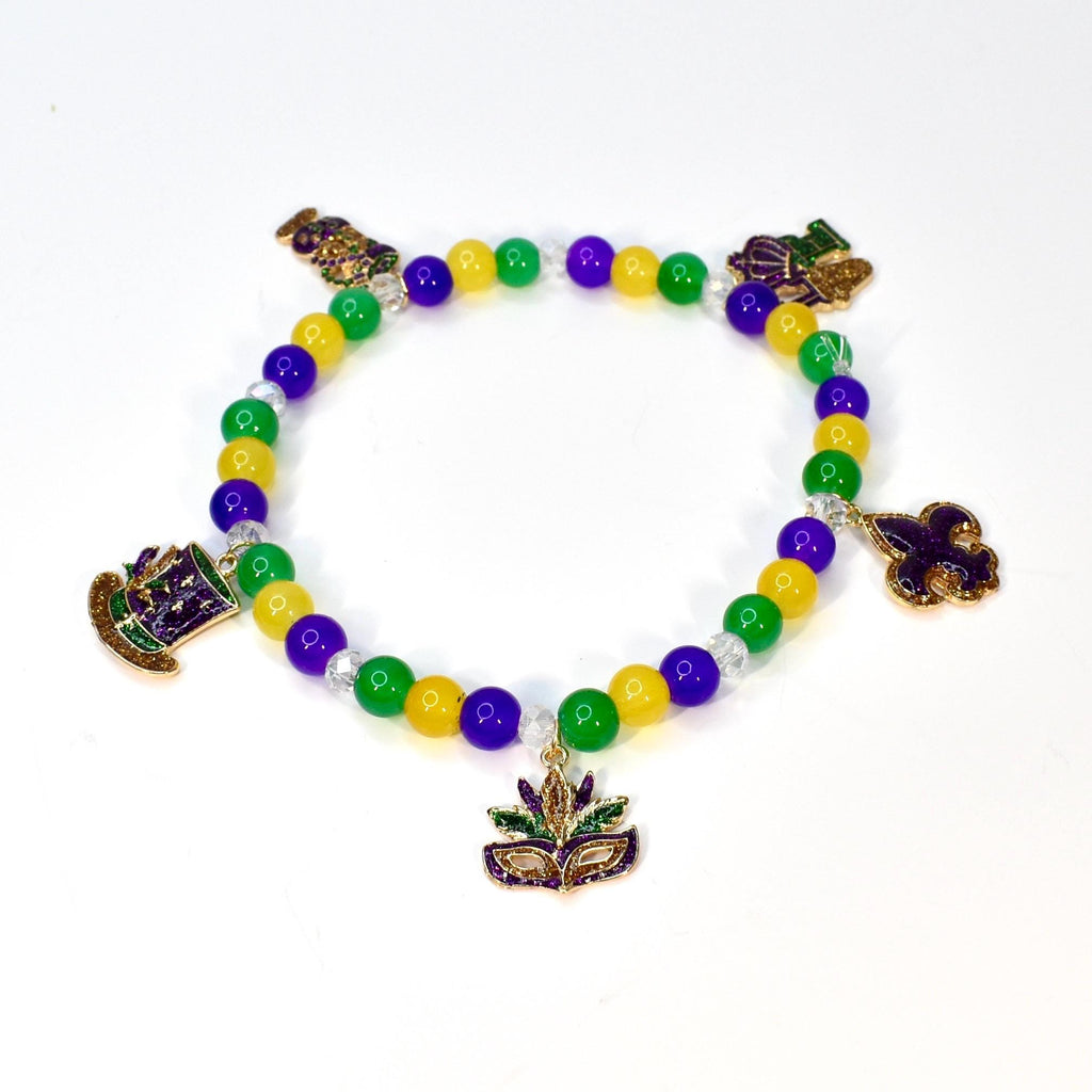 A beaded bracelet with yellow, green, and purple beads. The bracelet features gold-colored charms of a hat, a mask, and a fleur-de-lis. The beads are strung on a clear elastic cord, creating a festive accessory.