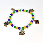 A beaded bracelet with yellow, green, and purple beads. The bracelet features gold-colored charms of a hat, a mask, and a fleur-de-lis. The beads are strung on a clear elastic cord, creating a festive accessory.